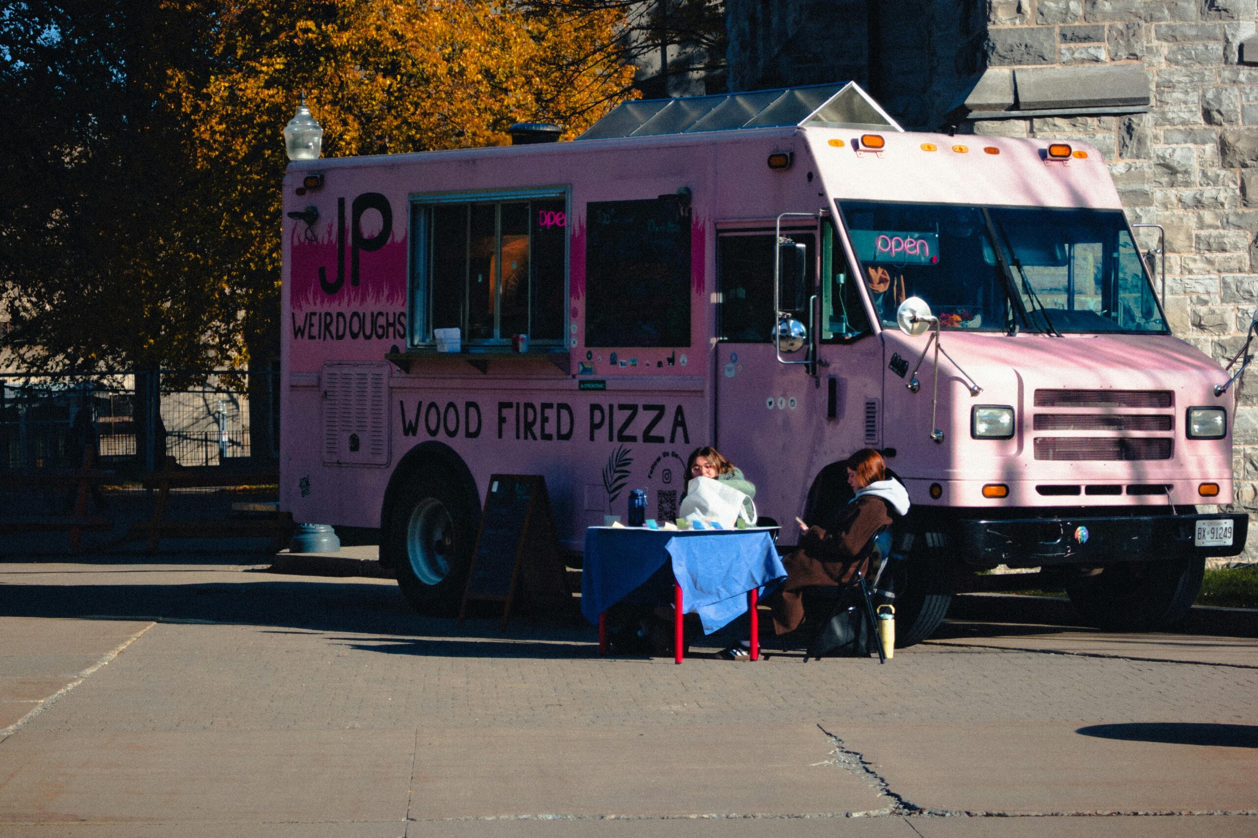 Colorful pink food truck serving wood-fired pizza on a sunny day in Kingston, Ontario.